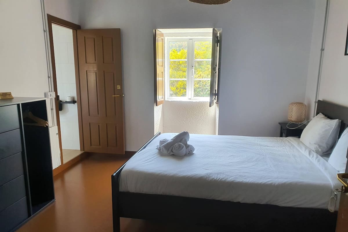 Una cama bien hecha con una toalla, una puerta de madera, una mesita con una lámpara y una ventana con vistas a árboles.