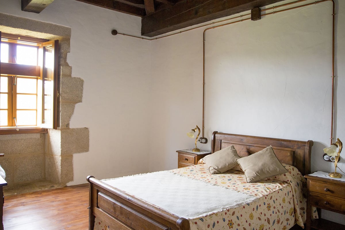 Un dormitorio con una cama de madera, una mesita de noche con lámpara, una ventana con cortinas y una pared de piedra.