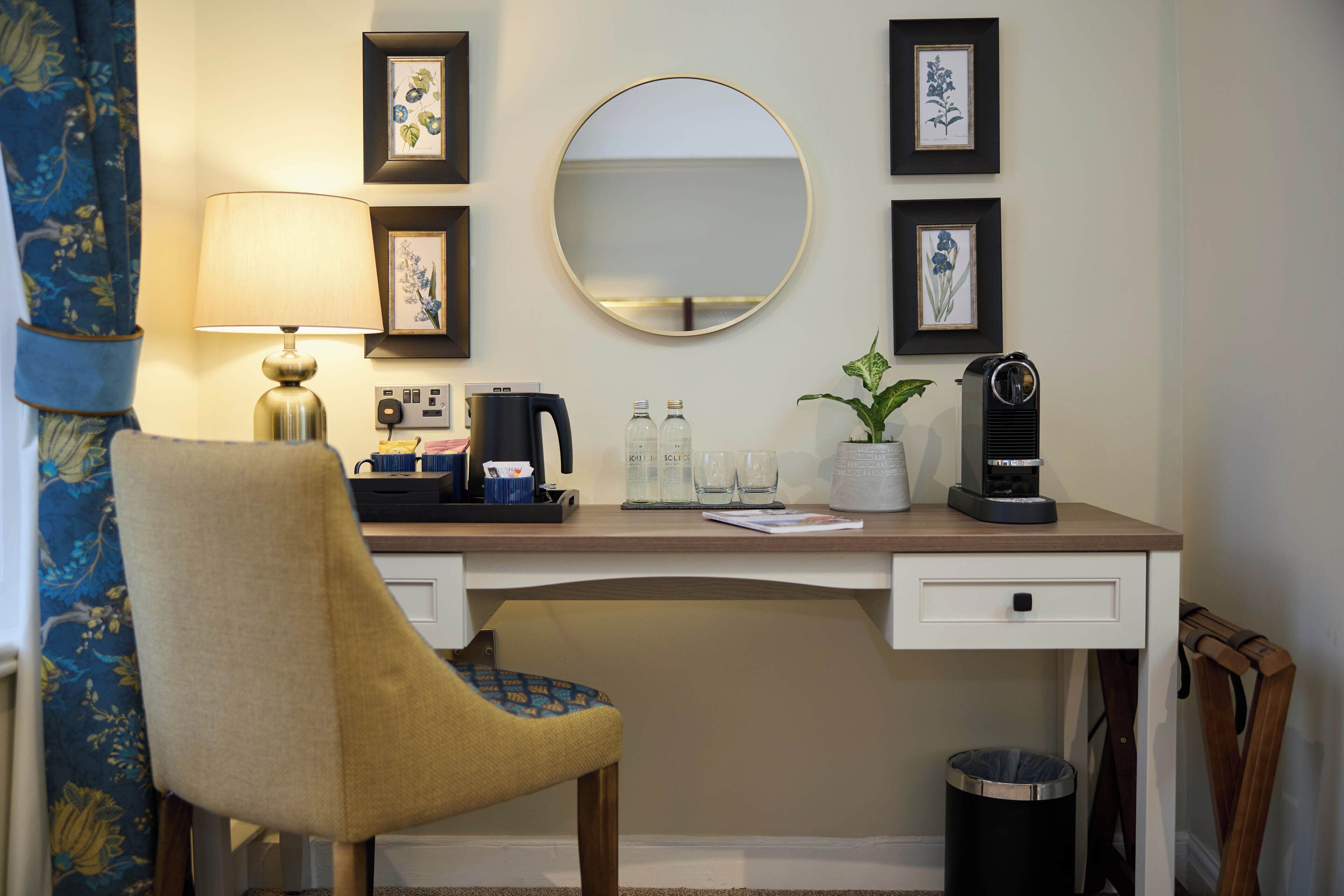 A hotel room with a desk, a chair, a lamp, framed pictures, a mirror, a coffee maker, and a trash can.