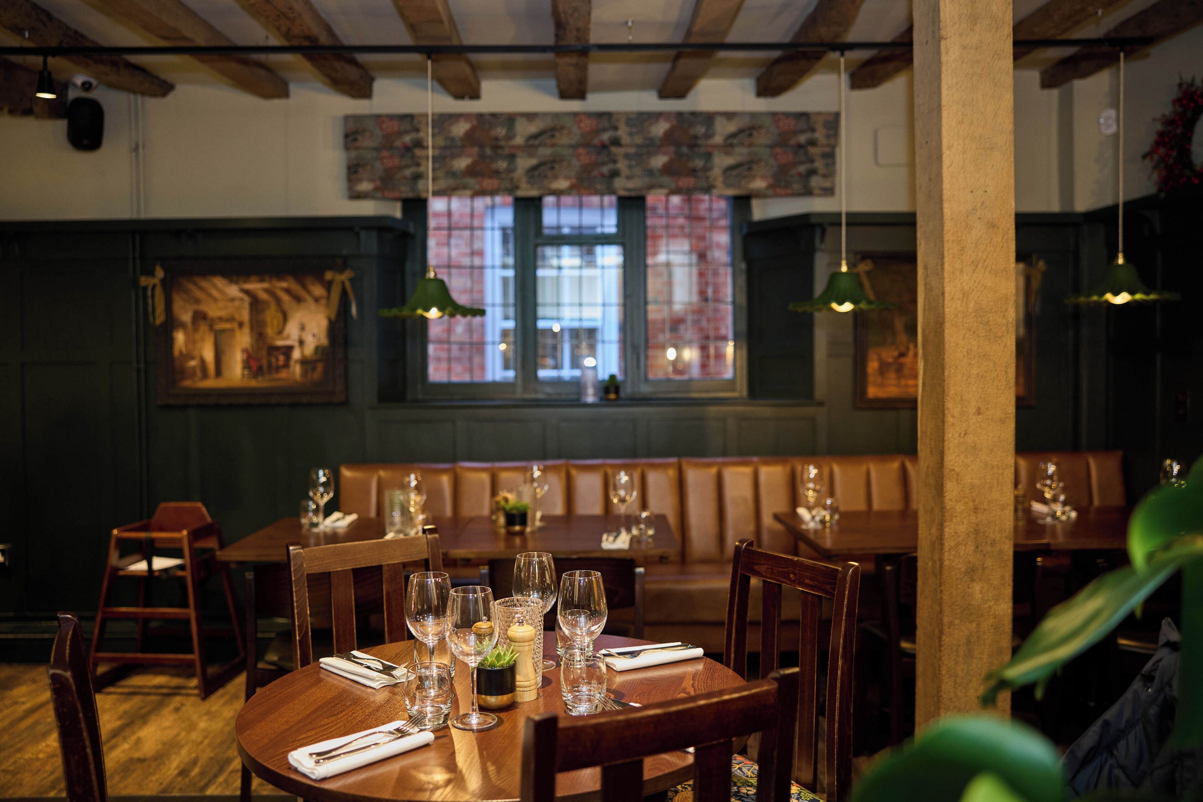 A restaurant interior with wooden tables, green hanging lamps, and a view through a window with red brick walls.