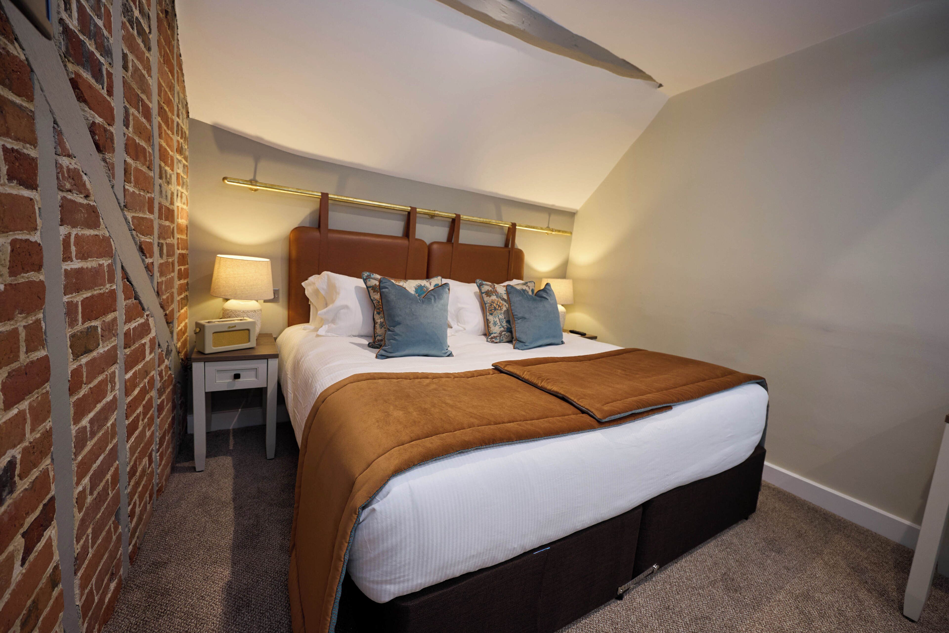 Une chambre avec un mur en briques, un lit avec une tête de lit marron, une table de chevet avec une lampe et un sol recouvert d’une moquette.