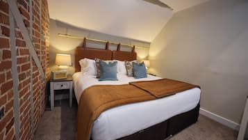 Une chambre avec un mur en briques, un lit avec une tête de lit marron, une table de chevet avec une lampe et un sol recouvert d’une moquette.