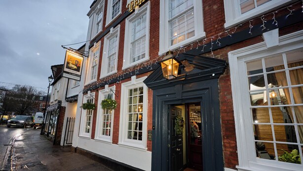 A traditional pub with a dark entrance door, white-framed windows, and hanging lights. - The White Hart (Bedford)
