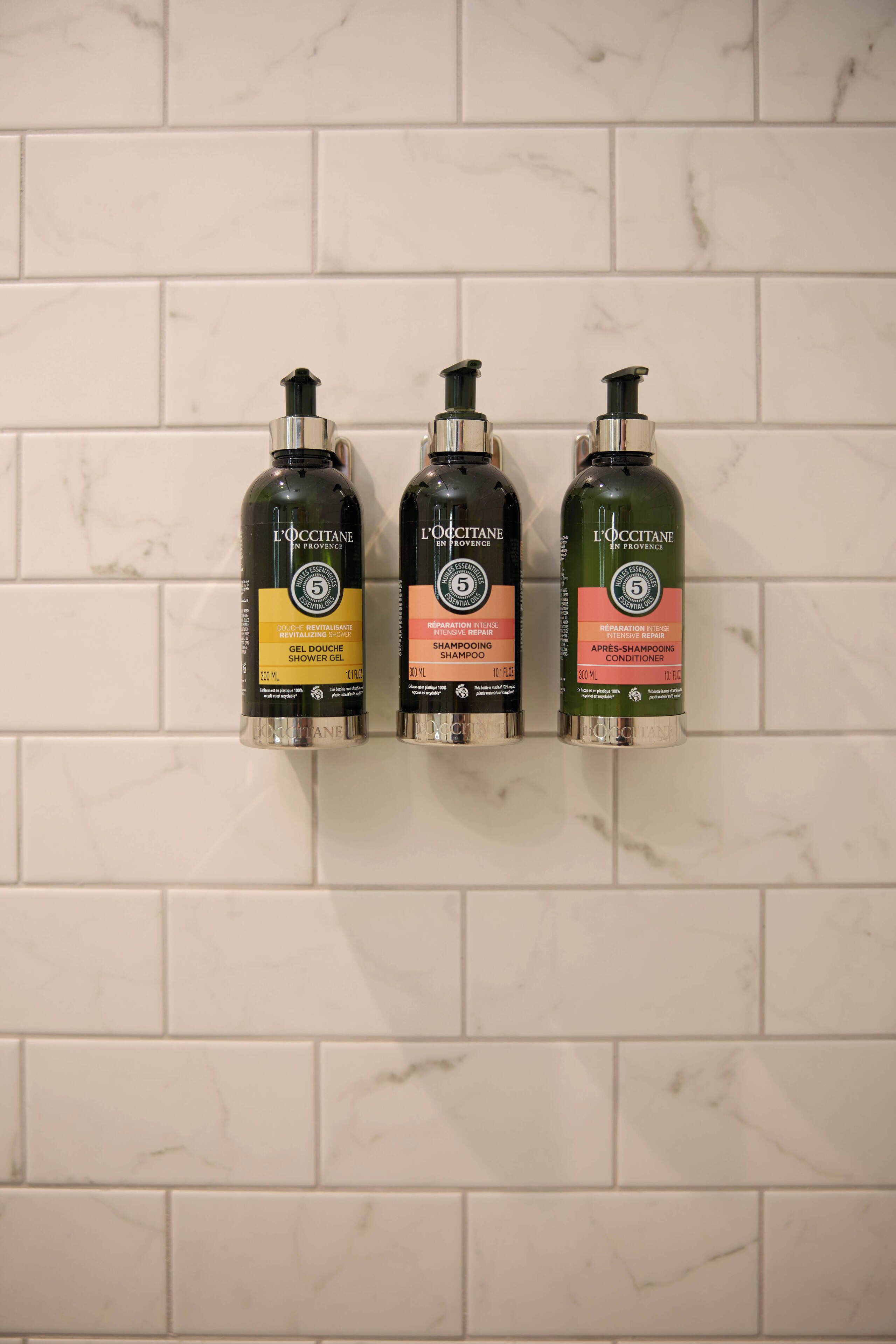 Tres botellas de gel de ducha L'OCCITANE sobre una pared de azulejos.