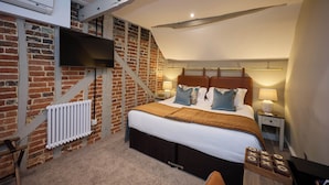A bedroom with a brick wall, a bed with a headboard, a TV mounted on the wall, a bedside table with a lamp, and a radiator. - The White Hart (Bedford)