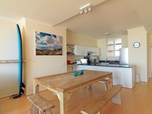 A kitchen with white cabinets, a wooden table, and a bench. There is a surfboard leaning against the wall and a painting of a landscape. - Surferscorner (Cape Town)