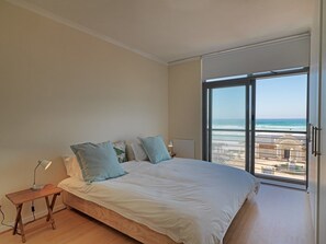 A bedroom with a large bed, a nightstand, and a view of the beach through a large window. - Surferscorner (Cape Town)