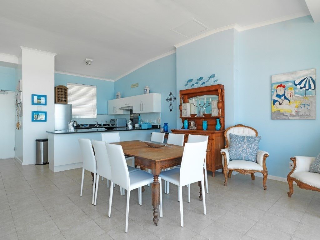 A dining area with a wooden table and white chairs, a kitchen with white cabinets, and a blue wall with artwork.
