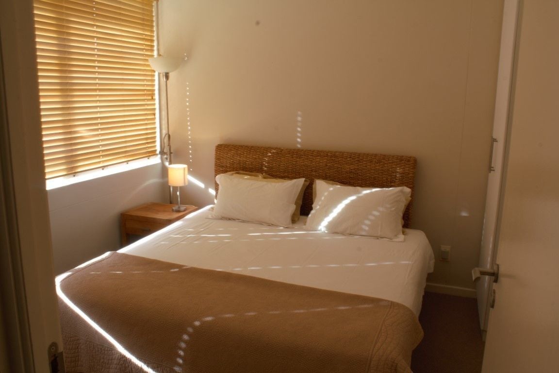 A neatly made bed with a brown headboard, a bedside lamp, and a window with blinds.