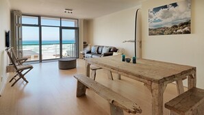 A spacious living area with a wooden table and benches, a leather sofa, and a large window offering a view of the ocean. - Surferscorner (Cape Town)