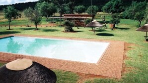 A rectangular swimming pool with a brick border, surrounded by a grassy area with trees and a wooden structure in the background.