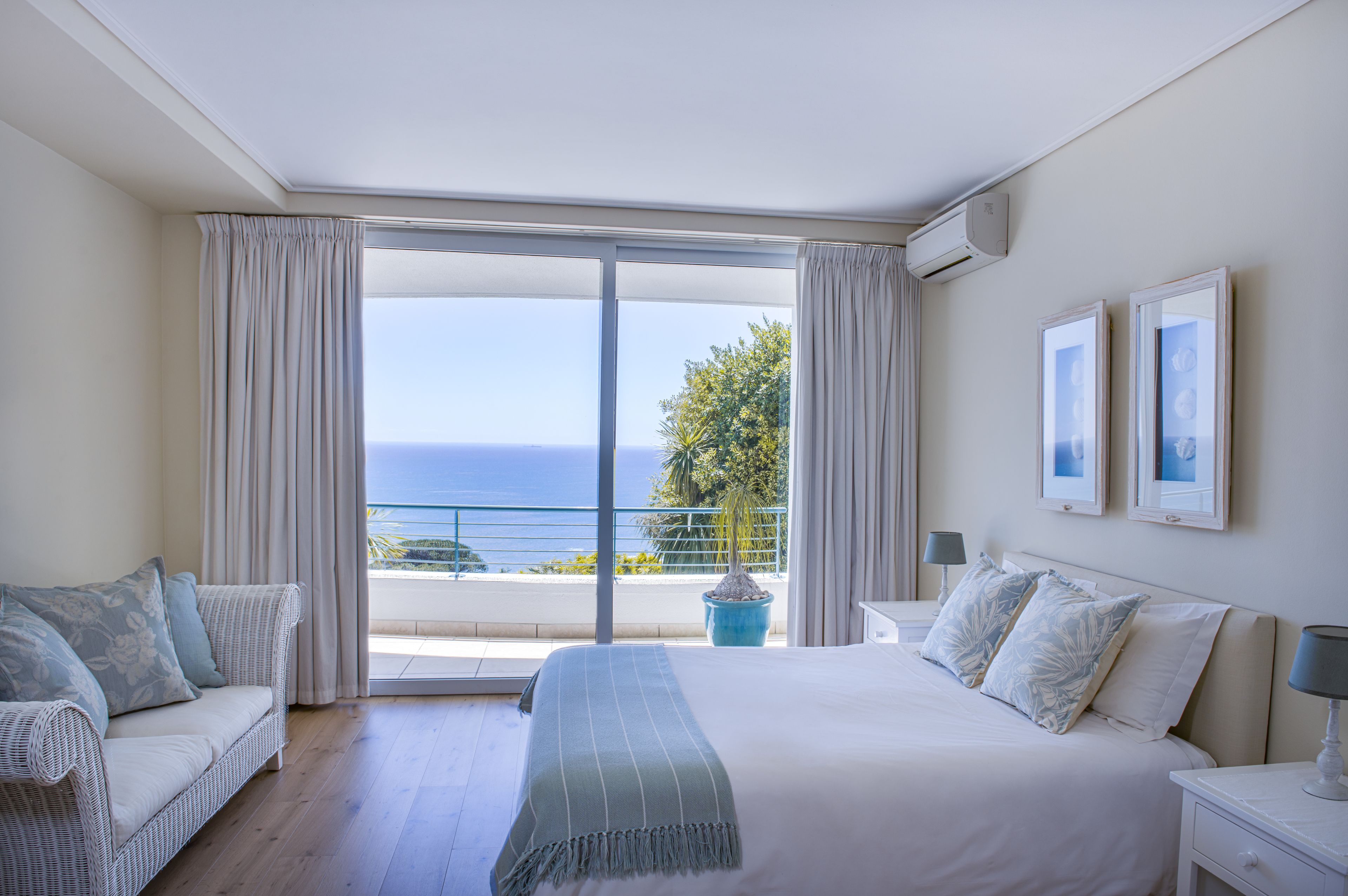 A hotel room with a large bed, a sofa, a balcony with a view of the sea, and two framed pictures on the wall.