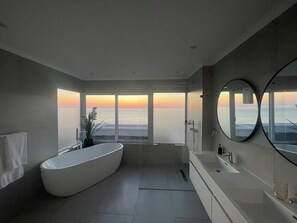 A modern bathroom with a large bathtub, a shower, and a view of the ocean at sunset.