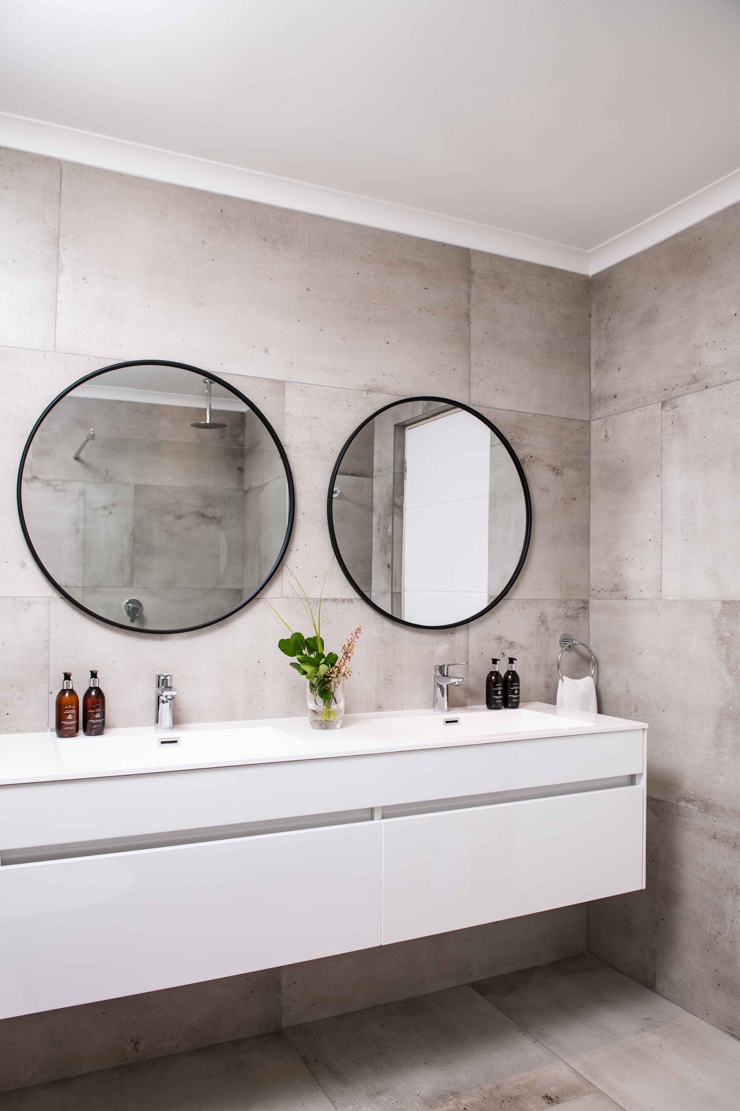 Ein modernes Badezimmer mit zwei runden Spiegeln, einem weißen Waschtisch und einer Betonwand.