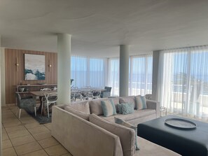 A modern living room with a beige sofa, a dining table, and a large window with sheer curtains.