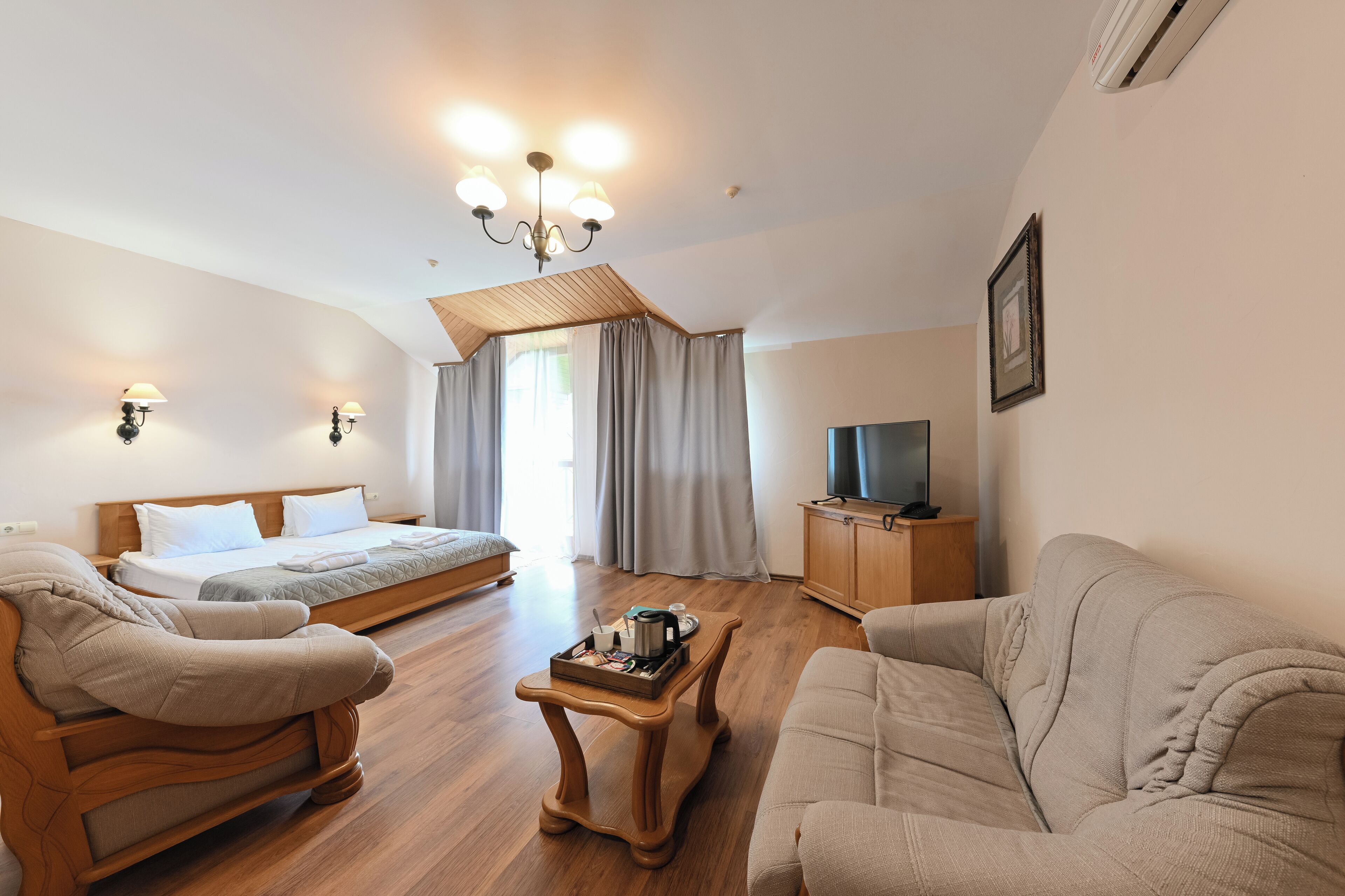 A well-lit room with a bed, two armchairs, a wooden coffee table, a television on a cabinet, and a chandelier.