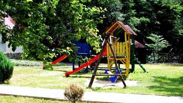 Children can play on the playground's wooden structure with a slide and climbing frame, surrounded by lush greenery and a tree-lined area.