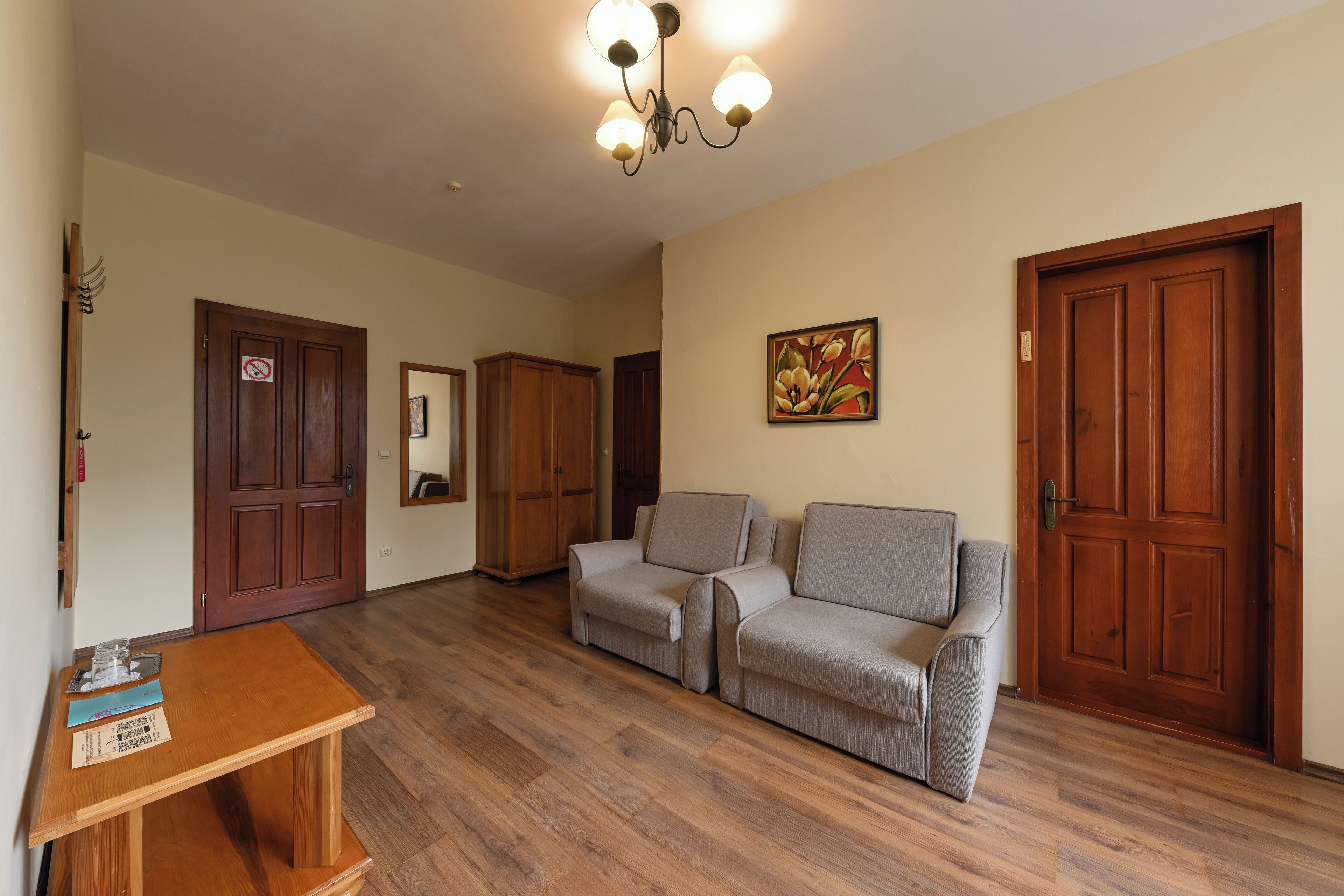 Un salón espacioso con suelo de madera, una mesa de madera con documentos, dos sofás grises, una puerta de madera con espejo y una araña.