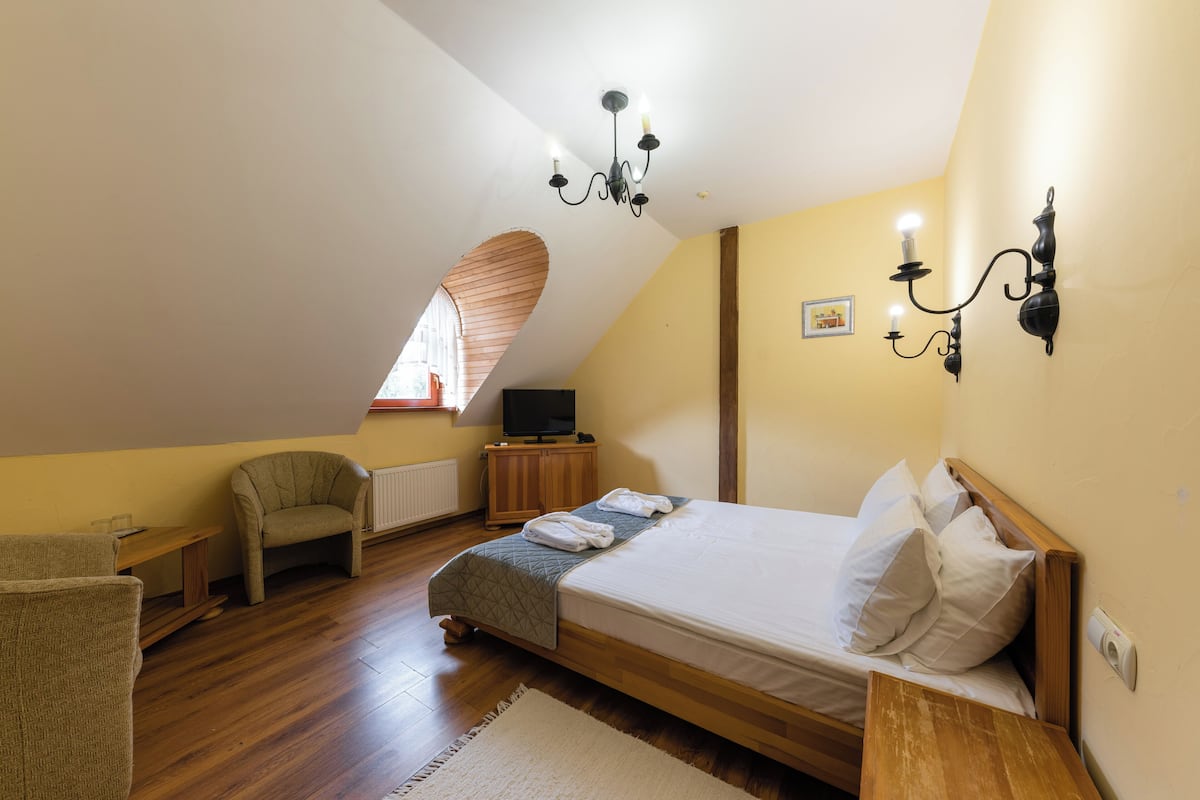 Un dormitorio con techo inclinado, una cama, una silla, un escritorio, un televisor y una lámpara fijada a la pared.
