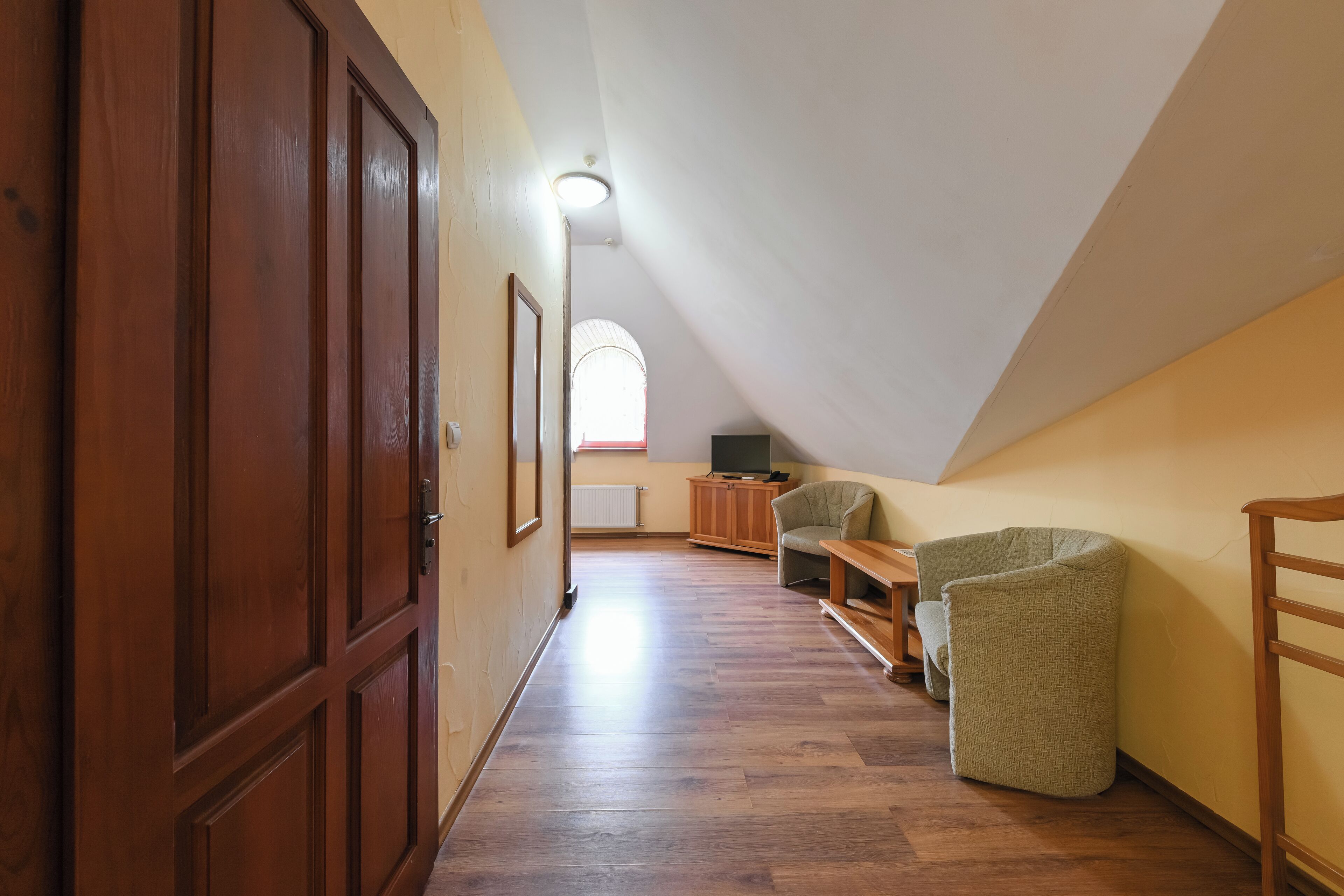 A narrow hallway with wooden flooring leading to a room with a sloped ceiling and a window.