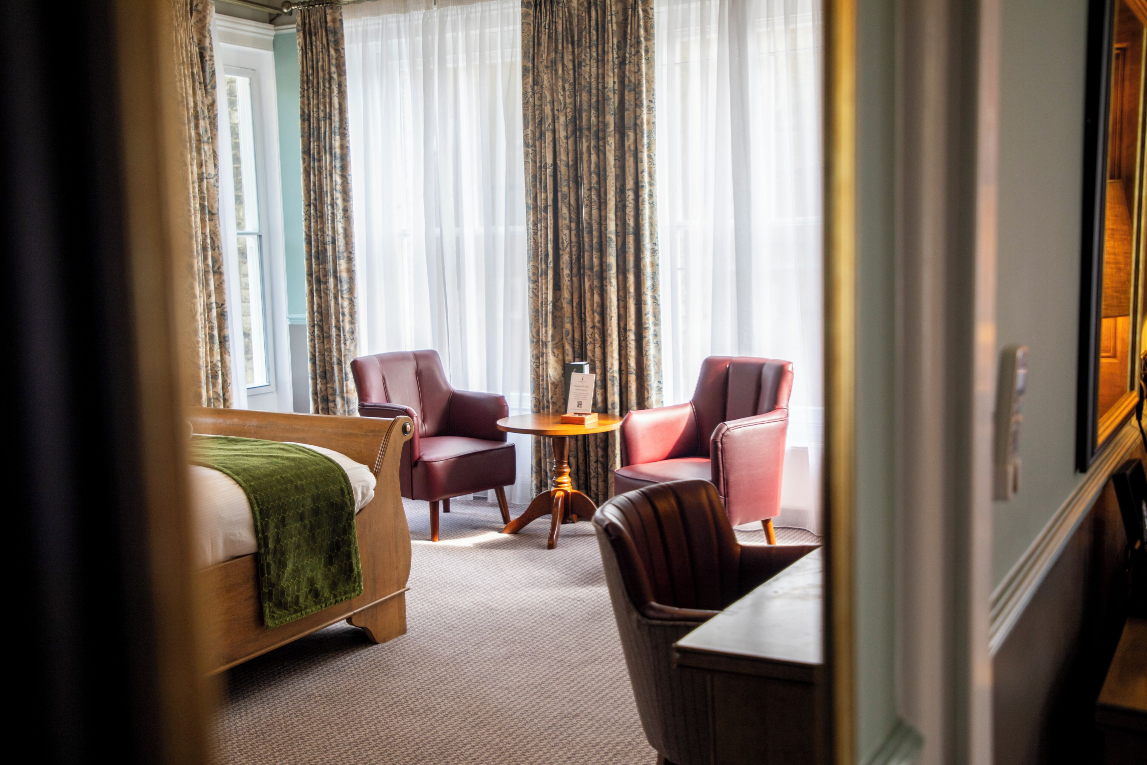 A hotel room with a bed, two leather armchairs, a small round table, and a view through sheer curtains.