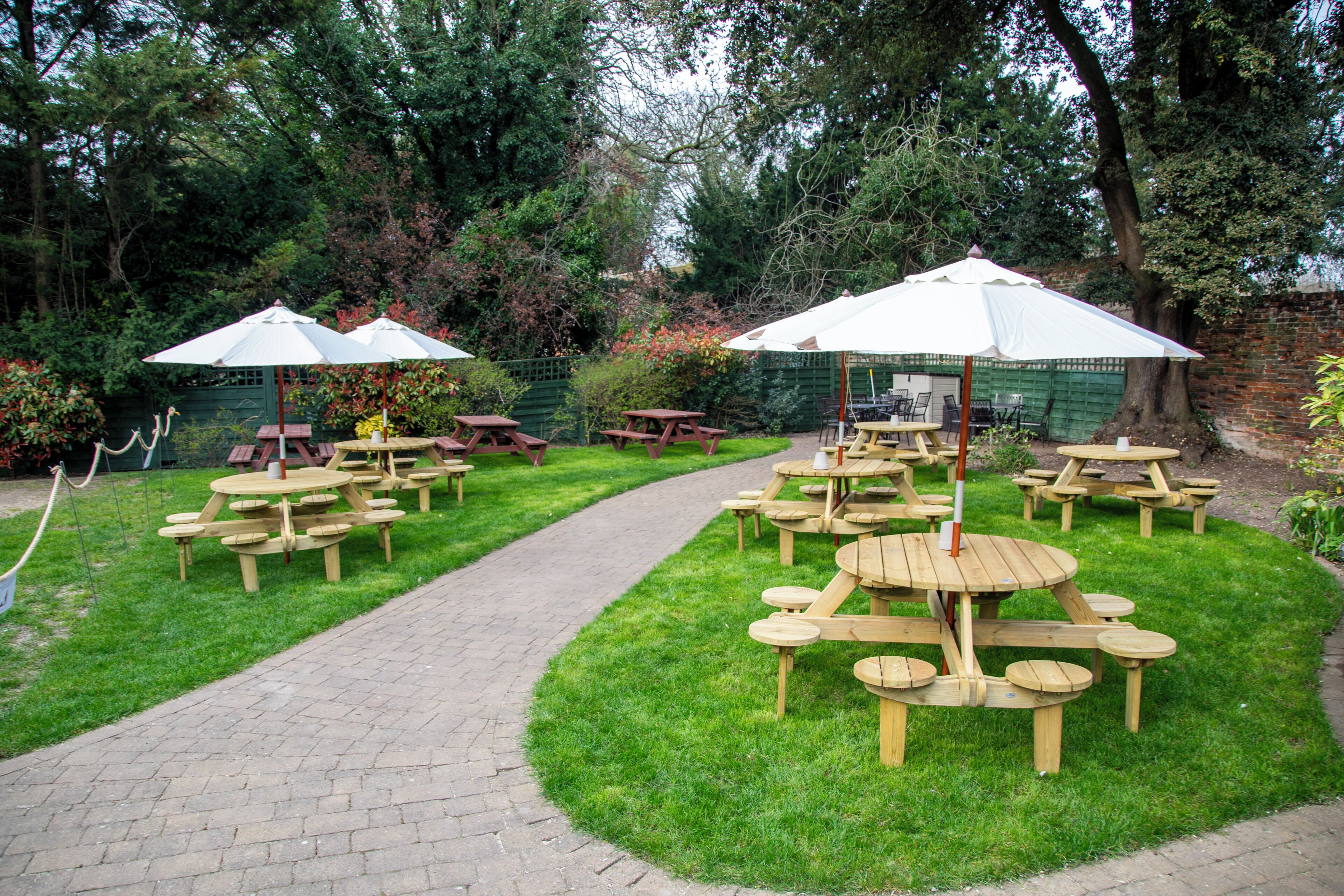 Un jardín con mesas de picnic y sombrillas, rodeado de árboles y un muro de ladrillo.