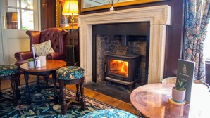 A room with a fireplace, leather armchair, two matching stools, a round table, and a mirror above the fireplace. - The Chalk Hare, Royston, Hertfordshire - Acorn Pubs (Royston)