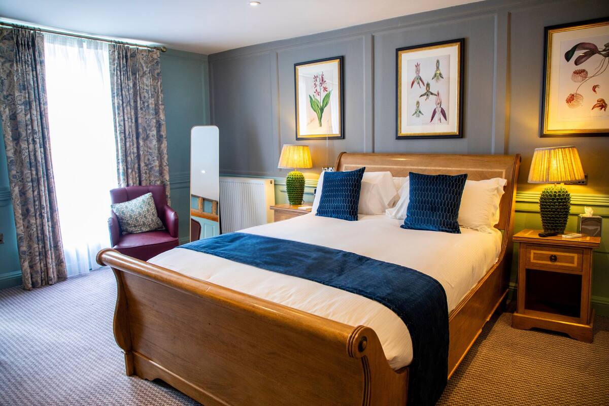 A hotel room with a large bed, a chair, two bedside tables with lamps, and framed botanical artwork on the walls.