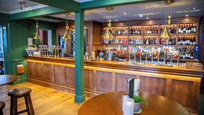 A bar with wooden shelves stocked with bottles, a counter with a cash register, and hanging pendant lights. - The Chalk Hare, Royston, Hertfordshire - Acorn Pubs (Royston)