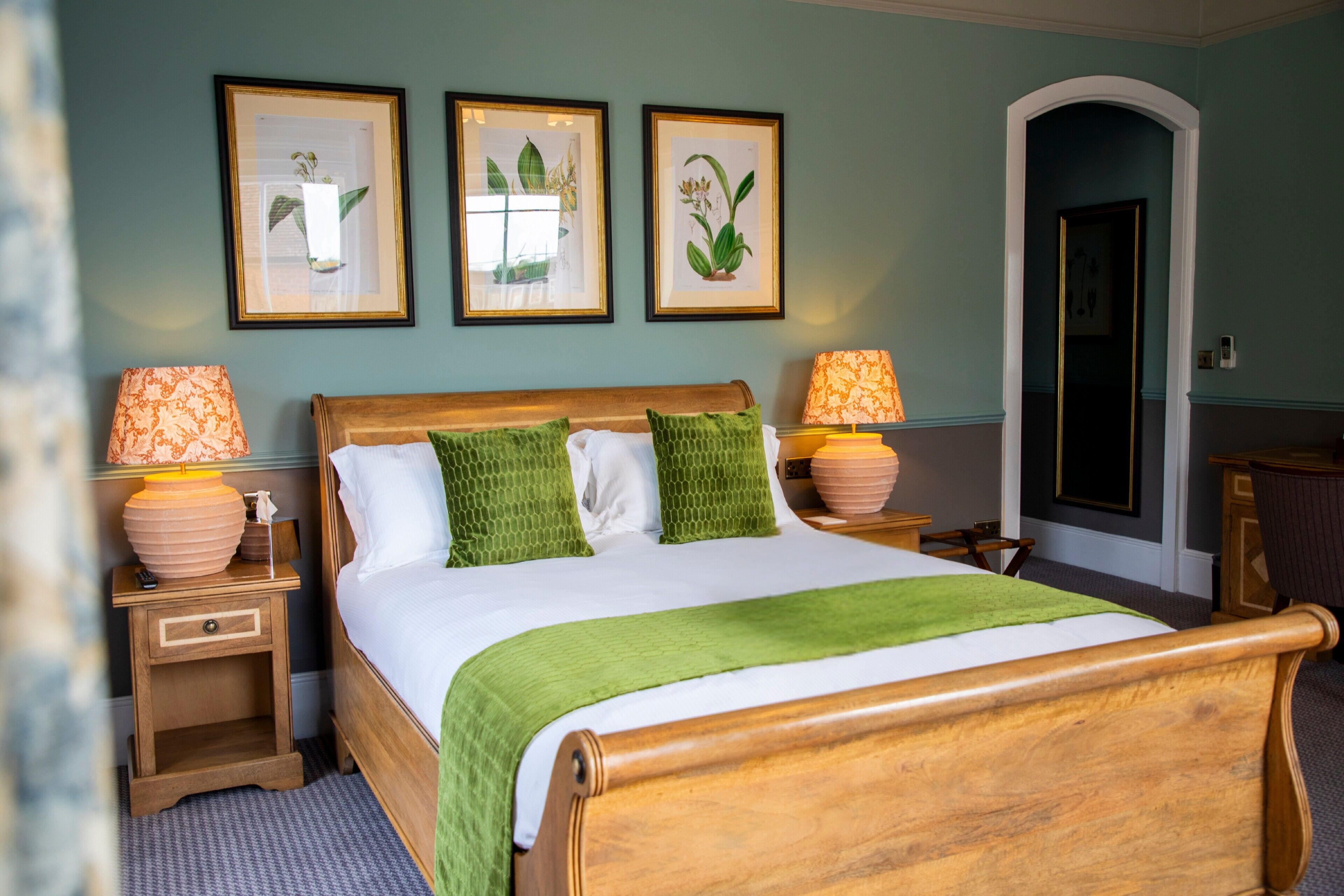 Un dormitorio ordenado con una cama de madera, almohadas verdes, dos lámparas de noche e impresiones botánicas enmarcadas en la pared.