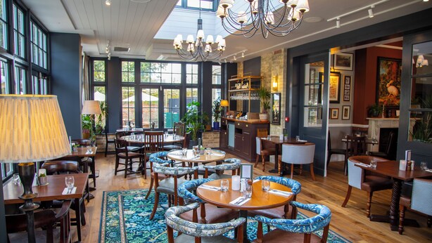 A spacious dining area with multiple tables and chairs, a chandelier, and large windows allowing natural light. - The Chalk Hare, Royston, Hertfordshire - Acorn Pubs (Royston)