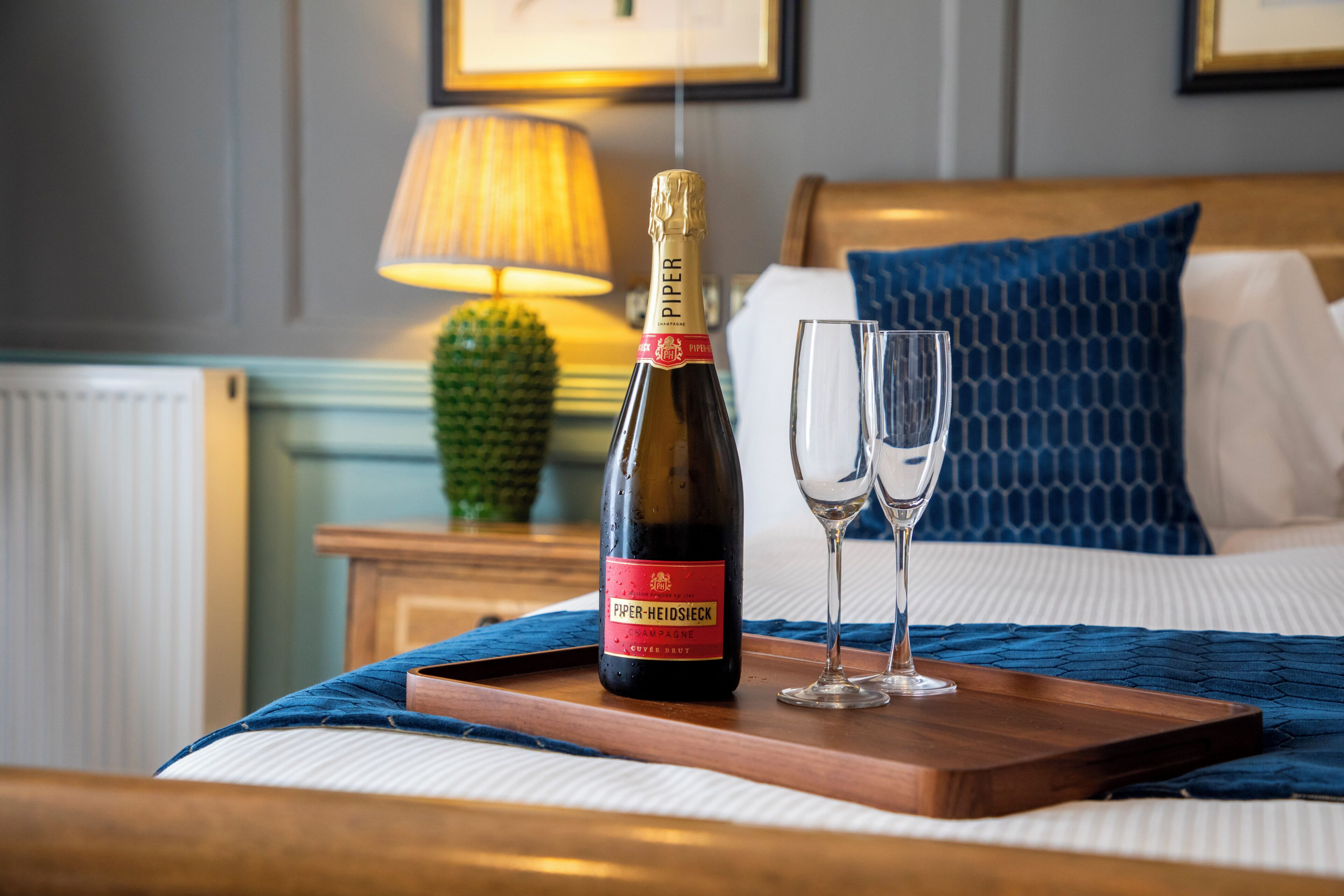A bottle of champagne and two glasses on a wooden tray on a bed with a blue blanket.