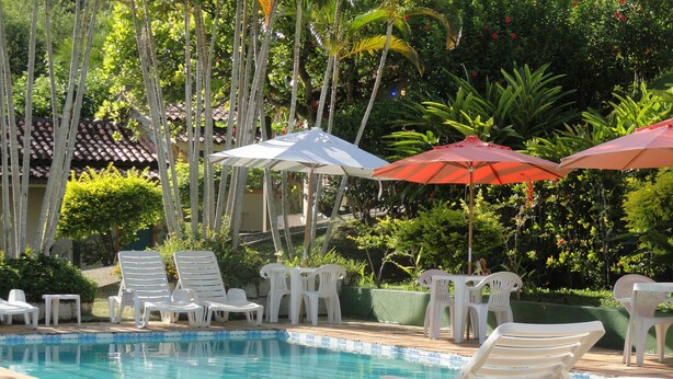 Uma área de piscina com espreguiçadeiras e mesas brancas, rodeada por vegetação e guarda-sóis.