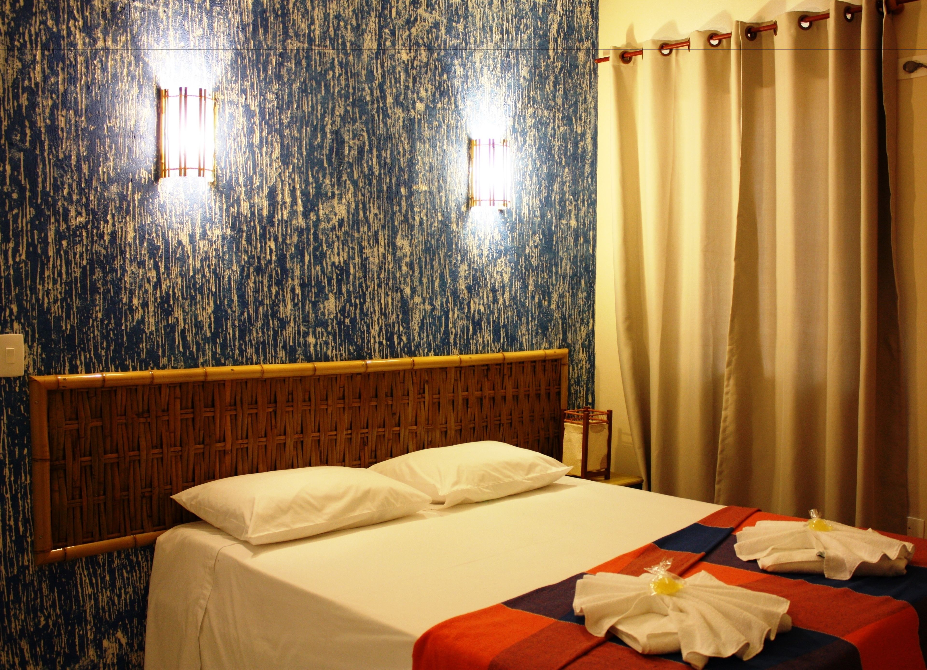 Una habitación de hotel con cama, cabecero, mesita de noche y cortina.