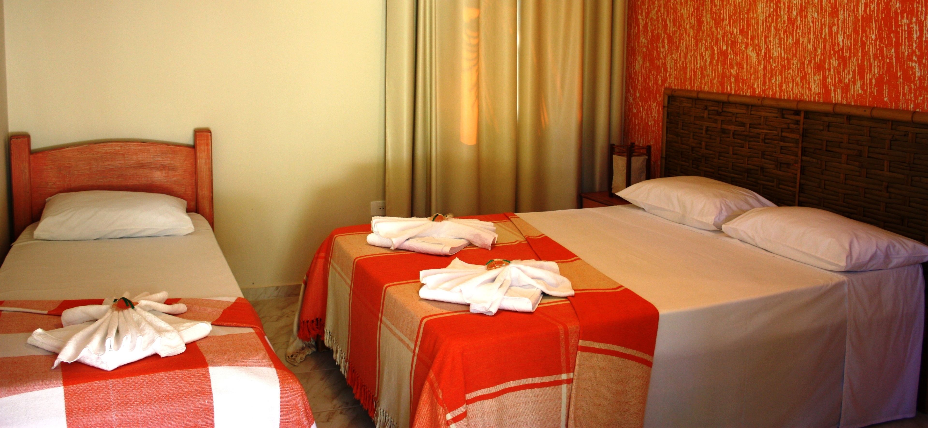 A hotel room with two single beds, each with a red and white checkered bedspread and a neatly folded white towel. The room has a wooden headboard and a red textured wall.