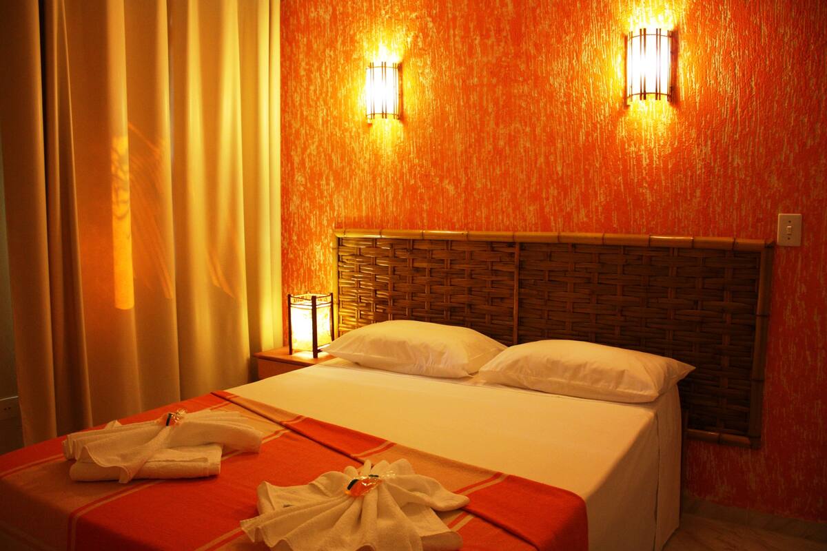 A hotel room with a bed, two bedside lamps, a woven headboard, and a red bedspread.