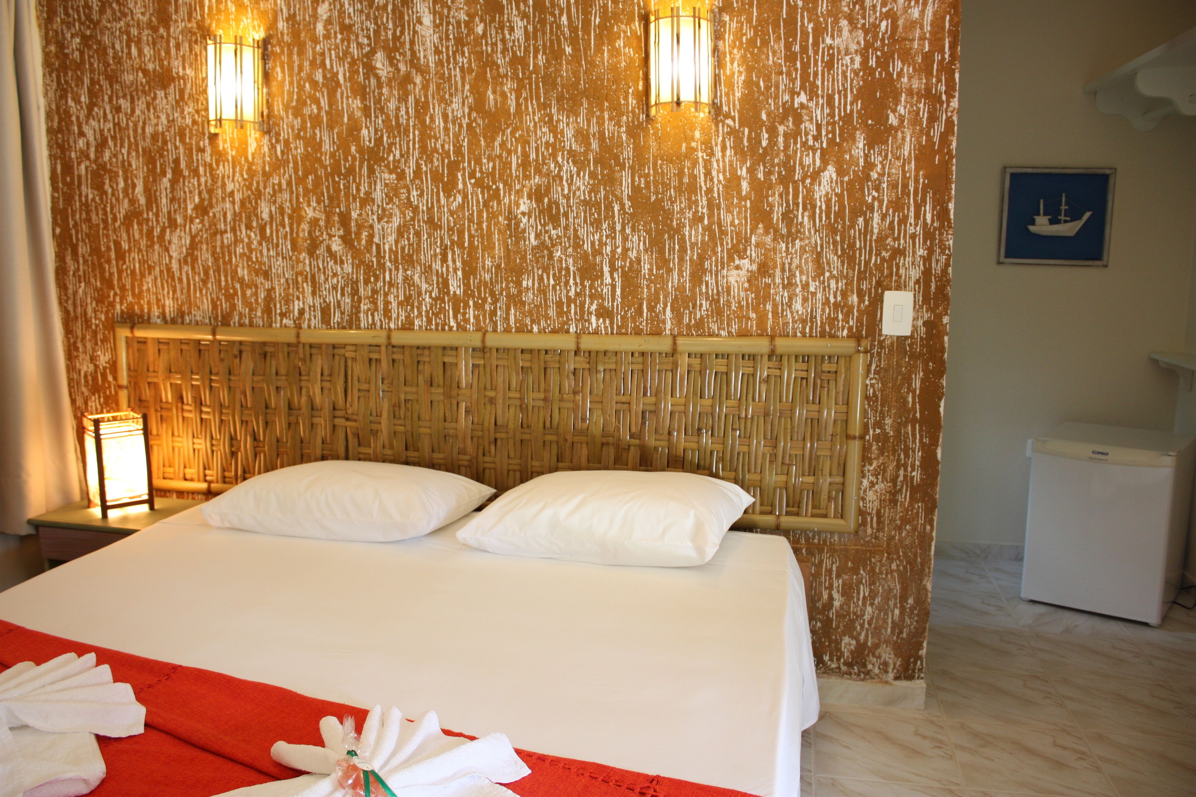A hotel room with a bed, two pillows, a red blanket, and a nightstand with a lamp.
