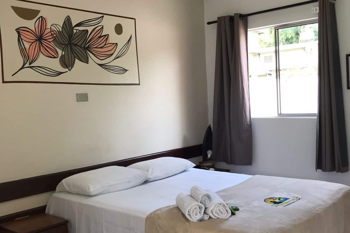 Ein Hotelzimmer mit Bett, Nachttisch, Deckenventilator, Fenster mit Vorhängen und einem Wandbild.