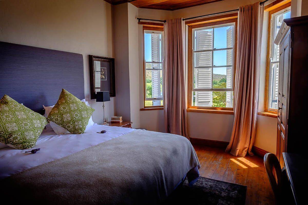 A bedroom with a bed, a nightstand, a lamp, a dresser, and large windows with curtains.