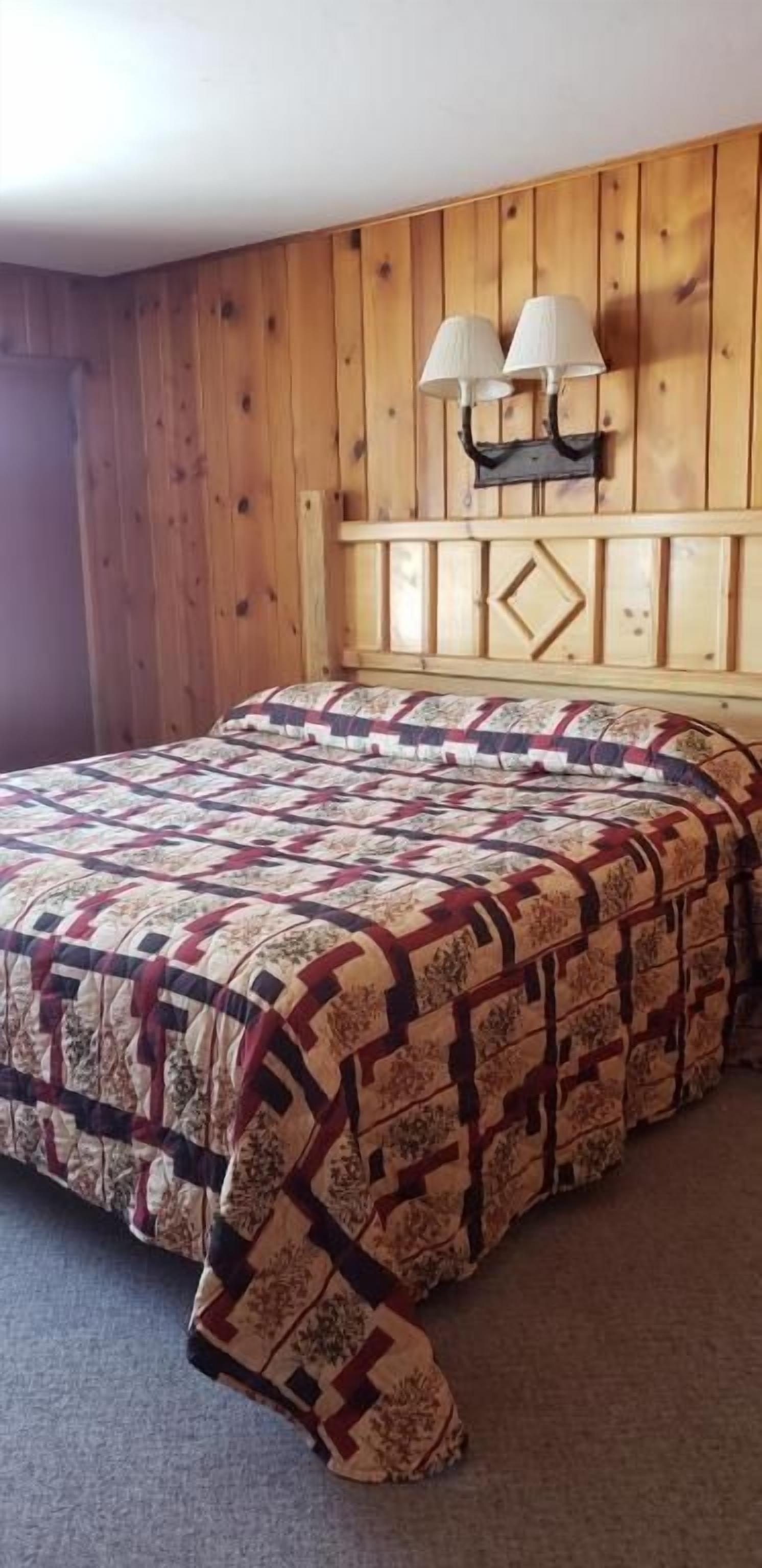 A bedroom with a wooden headboard, a bed with a patterned bedspread, and two wall-mounted lamps.