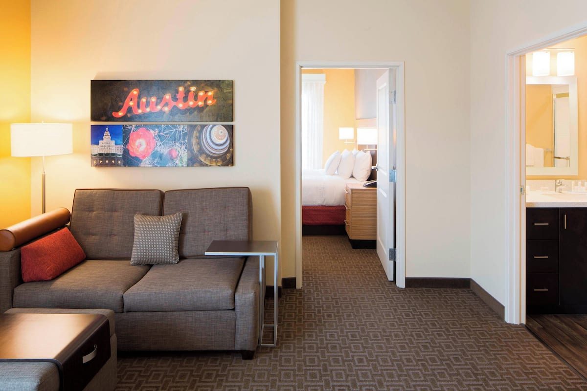 A hotel room with a grey sofa, a small table, a bed with white bedding, a bathroom with a mirror and sink, and a wall with a 'Austin' sign and artwork.