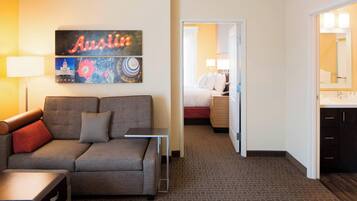 Una habitación de hotel con sofá gris, una mesita, cama con ropa de cama blanca, baño con espejo y lavabo, y una pared con un cartel de 'Austin' y obras de arte.