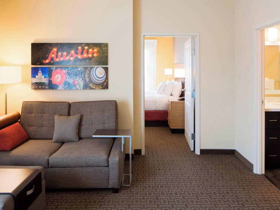 Una habitación de hotel con sofá gris, una mesita, cama con ropa de cama blanca, baño con espejo y lavabo, y una pared con un cartel de 'Austin' y obras de arte.