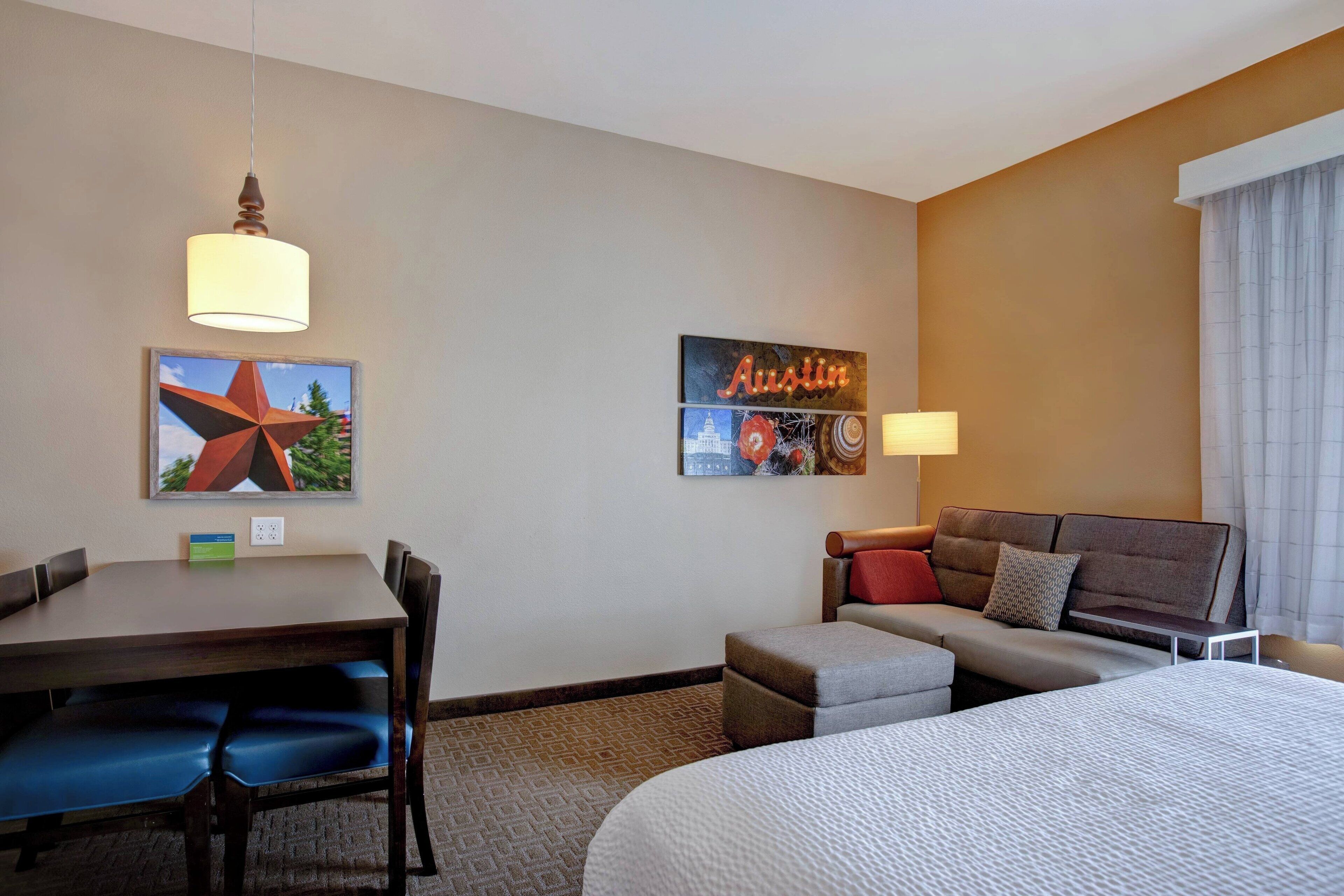 Quarto de hotel com cama, sofá, mesa de jantar com cadeiras e uma pequena mesa com abajur. Há decorações nas paredes e uma janela com cortinas.