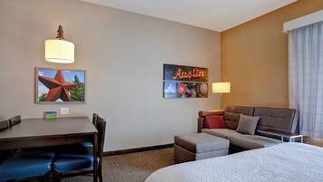 Quarto de hotel com cama, sofá, mesa de jantar com cadeiras e uma pequena mesa com abajur. Há decorações nas paredes e uma janela com cortinas.
