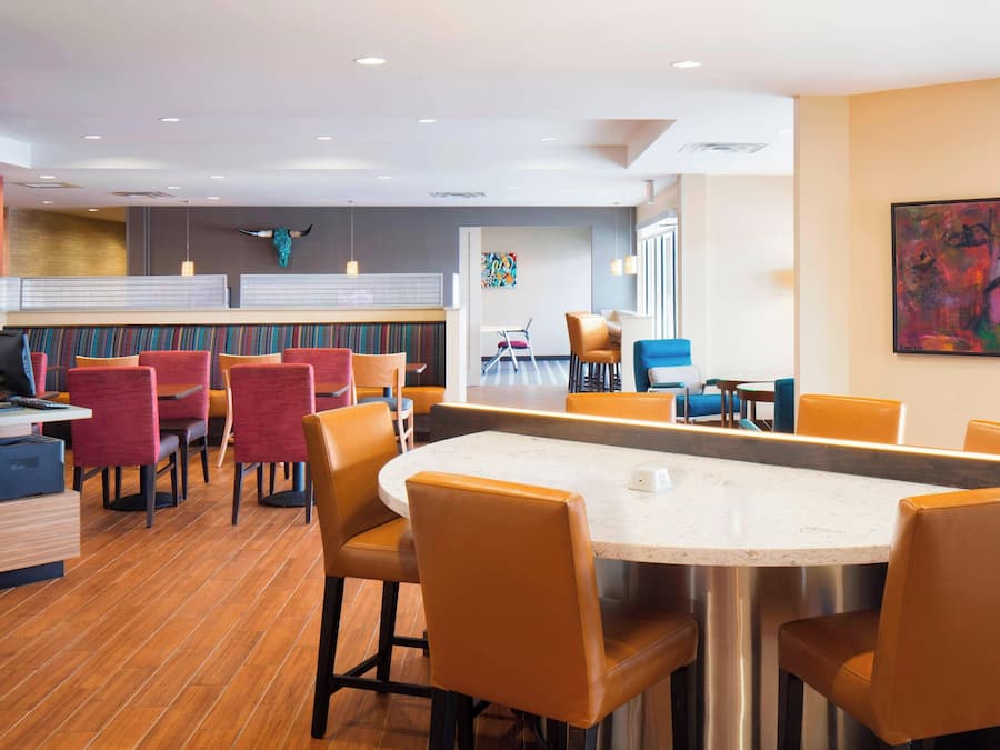 A modern hotel lobby with a central dining area featuring orange chairs and a marble table. There is a reception desk with a bull sculpture and a colorful abstract painting on the wall.