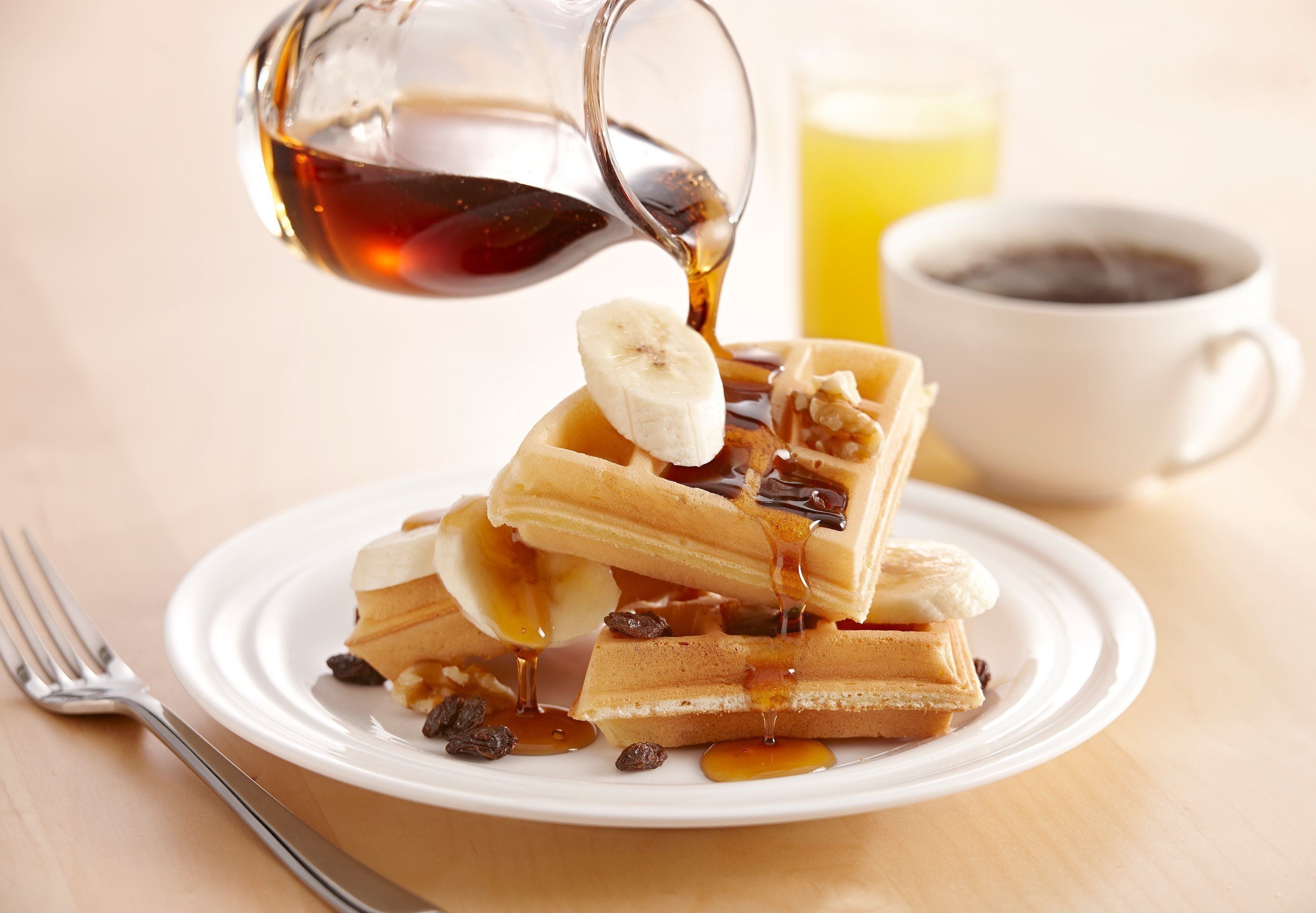 Un plato de gofres con plátanos y jarabe, un vaso de jugo de naranja y una taza de café sobre una mesa de madera.