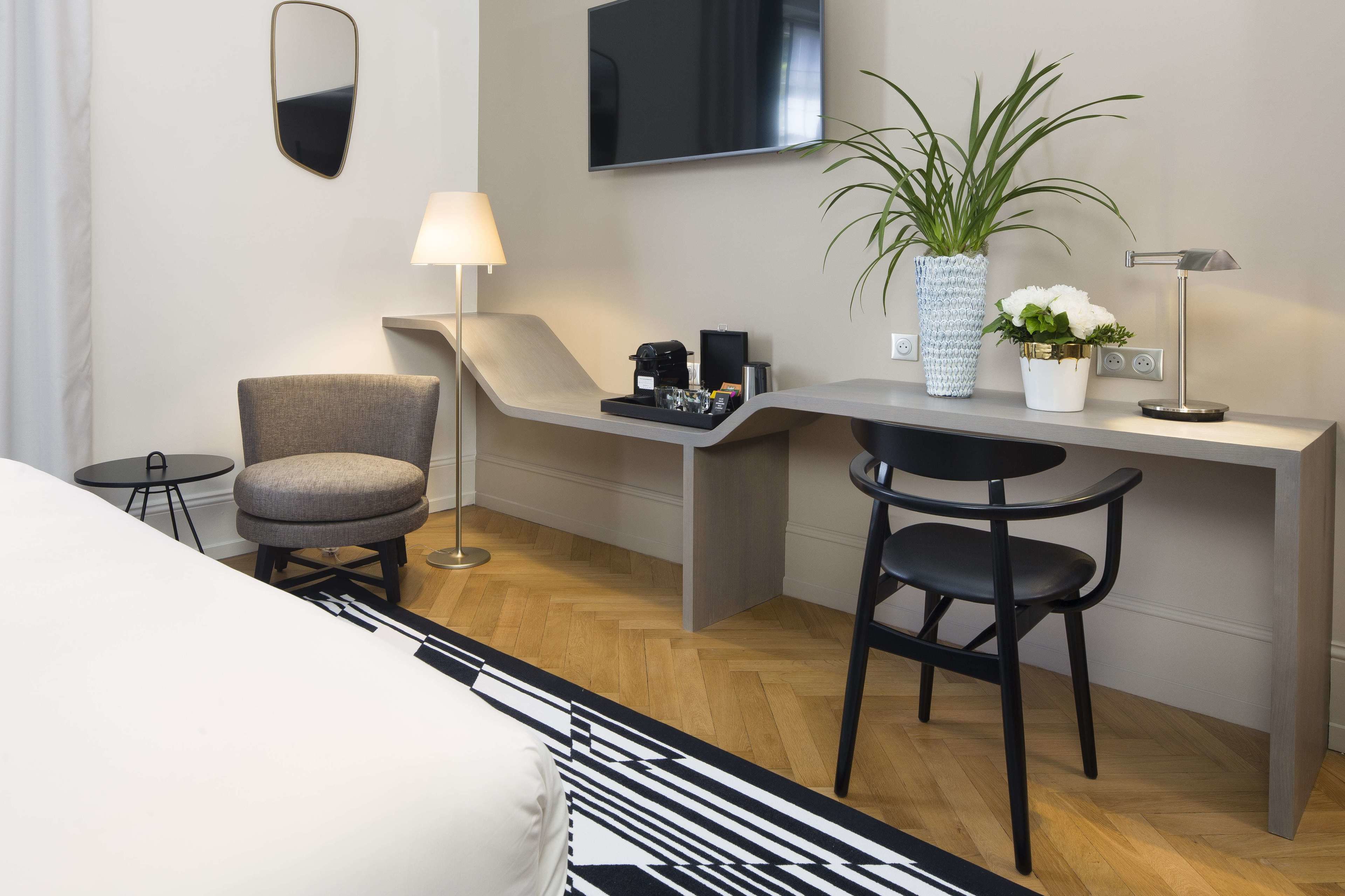 A modern hotel room with a desk, two chairs, a TV, a lamp, a potted plant, and a small table with a tray.