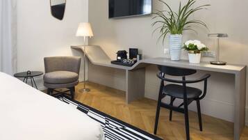 A modern hotel room with a desk, two chairs, a TV, a lamp, a potted plant, and a small table with a tray.