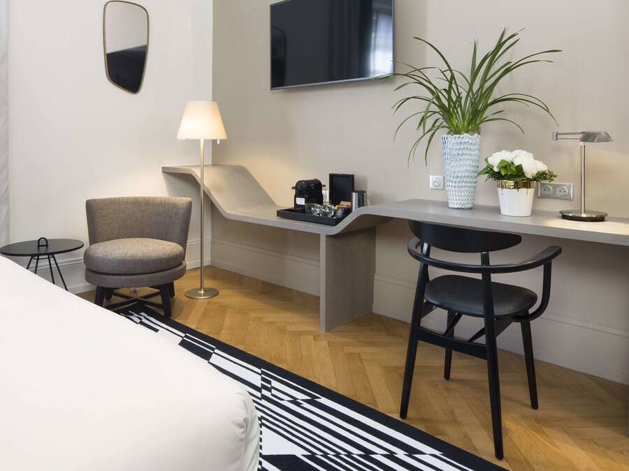 A modern hotel room with a desk, two chairs, a TV, a lamp, a potted plant, and a small table with a tray.